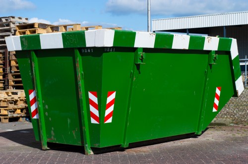 A large, rectangular green skip with reinforced metal edges and a slightly curved top, positioned on a paved surface in an outdoor setting. The skip features red and white reflective safety stripes on its sides, indicating it is used for waste collection or disposal. Behind the skip, there is a stack of wooden pallets on the left side and a blue industrial building with a corrugated roof in the background. The environment appears to be a commercial or industrial area, possibly near a waste management or recycling facility, with clear daylight illuminating the scene. The skip's sturdy construction and vibrant green colour denote it as part of a professional rubbish removal service, consistent with the offerings of Commercial Waste Removal Rotherhithe. The surroundings are neat, and the skip is ready for loading or pickup, reflecting the efficient waste collection operations in the area associated with local rubbish removal services.