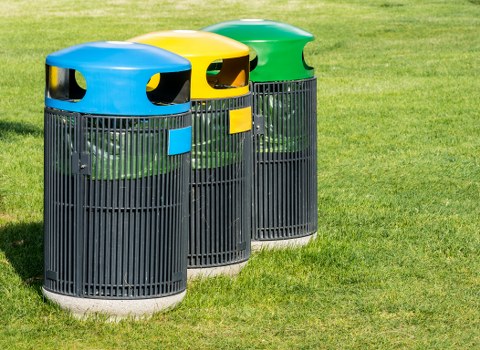 Three cylindrical outdoor rubbish bins are positioned side by side on a grassy area, with their openings facing forward. The bins are made of dark grey plastic with vertical slats, offering ventilation and visibility of their contents. Each bin has a differently coloured rounded plastic lid and matching rectangular label: one is blue, another yellow, and the third green. The grassy ground beneath is lush and well-maintained, and a blurred background suggests an outdoor open space, possibly near a residential or commercial building in Rotherhithe. The scene is evenly lit, indicating daylight conditions, and the focus is on the waste containers, representing waste management services such as those provided by Commercial Waste Removal Rotherhithe for general rubbish disposal.