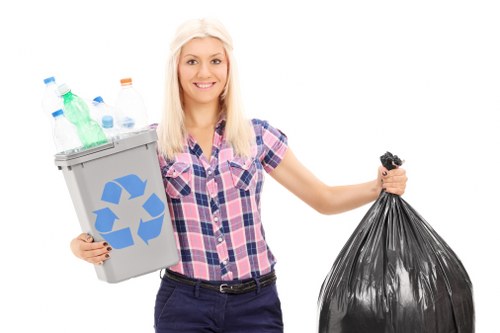 A young woman with long blonde hair, dressed in a casual short-sleeved, pink and blue checkered shirt and dark trousers, standing outdoors on a paved surface. She is smiling and holding a grey recycling bin filled with various plastic bottles, including clear, green, and blue caps, positioned in her left hand. In her right hand, she holds a large black plastic rubbish bag, tied at the top. The background is plain white, emphasizing the waste items and her cheerful expression, illustrating a rubbish removal or recycling concept. The scene emphasizes responsible waste disposal and aligns with services such as those offered by Commercial Waste Removal Rotherhithe.
