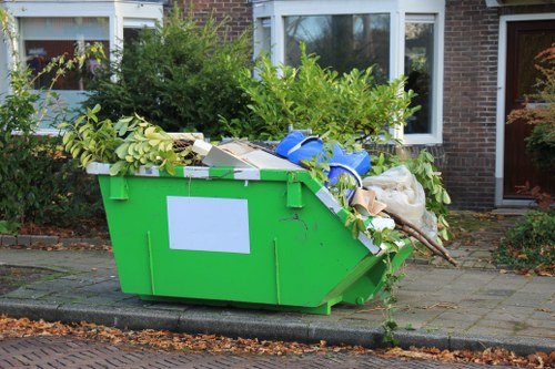 A bright green, large rectangular waste skip situated on a paved driveway in front of a residential brick house with white-framed windows. The skip is filled with various types of rubbish, including green leafy branches, cardboard boxes, plastic bags, and miscellaneous discarded items, with some materials spilling over the top edge. The surrounding area features a garden with hedges and small trees, with the skip positioned close to the pavement and curb. The background shows the front of the house with a slightly overcast sky, and the scene appears to be a typical suburban setting, possibly in Rotherhithe. The image illustrates an outdoor rubbish collection point ready for waste removal services, such as those provided by Commercial Waste Removal Rotherhithe.