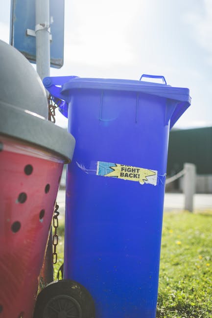 An outdoor scene showing a pile of mixed rubbish and waste materials situated on a gravel surface. The waste includes several black plastic garbage bags, some partially torn open, revealing contents s